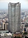Aerial view of one curved side of a tall building set in front of a cityscape; the facade is symmetrical and divided into three vertical sections: both identical sections on either side are divided by a section in the center that turns into a large atrium window as it nears the building's roof