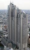 Aerial view of a building's two beige facades with horizontal rows of windows set in front of a cityscape; the building is composed of three adjoined towers of differing heights, each capped off with triangular glass structures