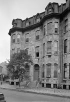 End section of a four-story stone terraced building. Two adjacent doors are at center, each with a short flight of stairs leading up from the street to the first level. Either side of the doors, at left and right, the building has two rounded tower-like sections, each with nine large sash windows (three on each level). Four more large windows are at center, above the doors. The uppermost (attic) level has four smaller windows protruding from the roof with architectural feature. At street level, grilled skylights illuminate basement rooms.