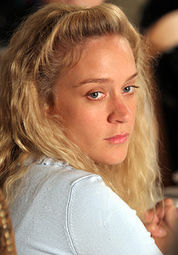 Blonde, wavy-haired woman with a very solemn expression looking over her shoulder.
