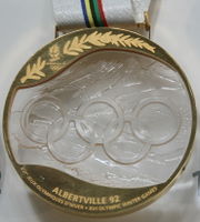 A crystal inscribed with five rings and a mountainous background, surrounded by gold making a complete medallion.  The gold is inscribed with a branch and the logo for these Games at the top, with the bottom containing the words "ALBERTVILLE 92; XVIth Jeux Olympiques D'Hiever; XVI Winter Olympic Games".