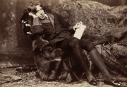 A tall man rests on a chaise longue, facing the camera. On his knees, which are held together, he holds a slim, richly bound book. He is wearing knee breeches which feature prominently in the photograph's foreground. Oscar Wilde reclines with Poems for Napoleon Sarony in New York in 1882. Wilde often liked to appear idle, though in fact he worked hard; by the late '80s he was a father, an editor, and a writer.