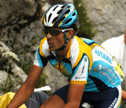 A cyclist in a blue and yellow jersey with white trim. His bicycle is not visible, but he is in riding position. He wears sunglasses, a helmet to match his jersey, and a bandage on his nose.