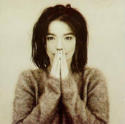 A picture of the album cover depicting a muted background with Björk standing facing forward in the middle. Björk is dressed in a fuzzy ragged sweater with her hands close together covering most of her mouth.