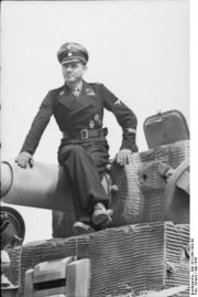 A man, wearing dress uniform and a cap, sits on top of a tank barrel; the tank is not fully in view.