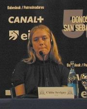 Head and bust profile of slender woman sitting at table, clad in a dark blouse, with long blonde hair. A small microphone is perched in front her.