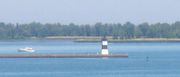 A pier with a square, black and white-striped lighthouse at the end. A small motorboat is paralleling the pier, heading away from the lighthouse.
