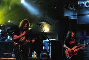 Two men with long hair are on a stage.  One plays a shiny guitar and the other plays a bass guitar with a wood finish