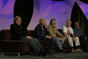 Five older men sit on a couch, with an arched, decorative purple background behind them. Ralston, leaning back in his seat, has short hair and wears a shirt with slacks.