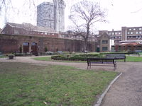 A grassy garden with some flower beds, bushes, two bare trees, walkways and park benches. It is bounded on its left side by a 15-foot-high brick wall, with a closed wrought-iron double-arched gateway in the wall. On the far side of the garden is a little two-story off-white house with green windows, a peaked roof, and a door into the garden. In the far background can be seen the tops of several apartment complexes and a 20-story office building.