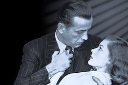 Black-and-white image of a man and a woman, seen from mid-chest up, their faces in profile, gazing into each others' eyes. He embraces her in a dip with his right arm and holds her right hand to his chest with his left hand. He wears a pin-striped suit and a dark tie. She wears a white top. On the left, the background is black; on the right, it is lighter, with a series of diagonal shadows descending from the upper corner.
