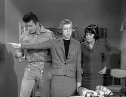 A black and white screenshot from the television series, The Beverly Hillbillies shows Max Baer Jr. as Jethro, Nancy Kulp as Jane Hathaway and Sharon Tate as Janet Trego, a secretary. Tate is wearing a business suit and a dark wig and is watching Miss Hathaway