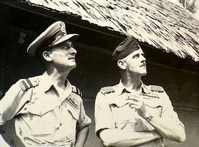 Half-length informal portrait of two men in short-sleeved tropical military uniforms with pilot's wings, standing in front of a hut and looking into the sky